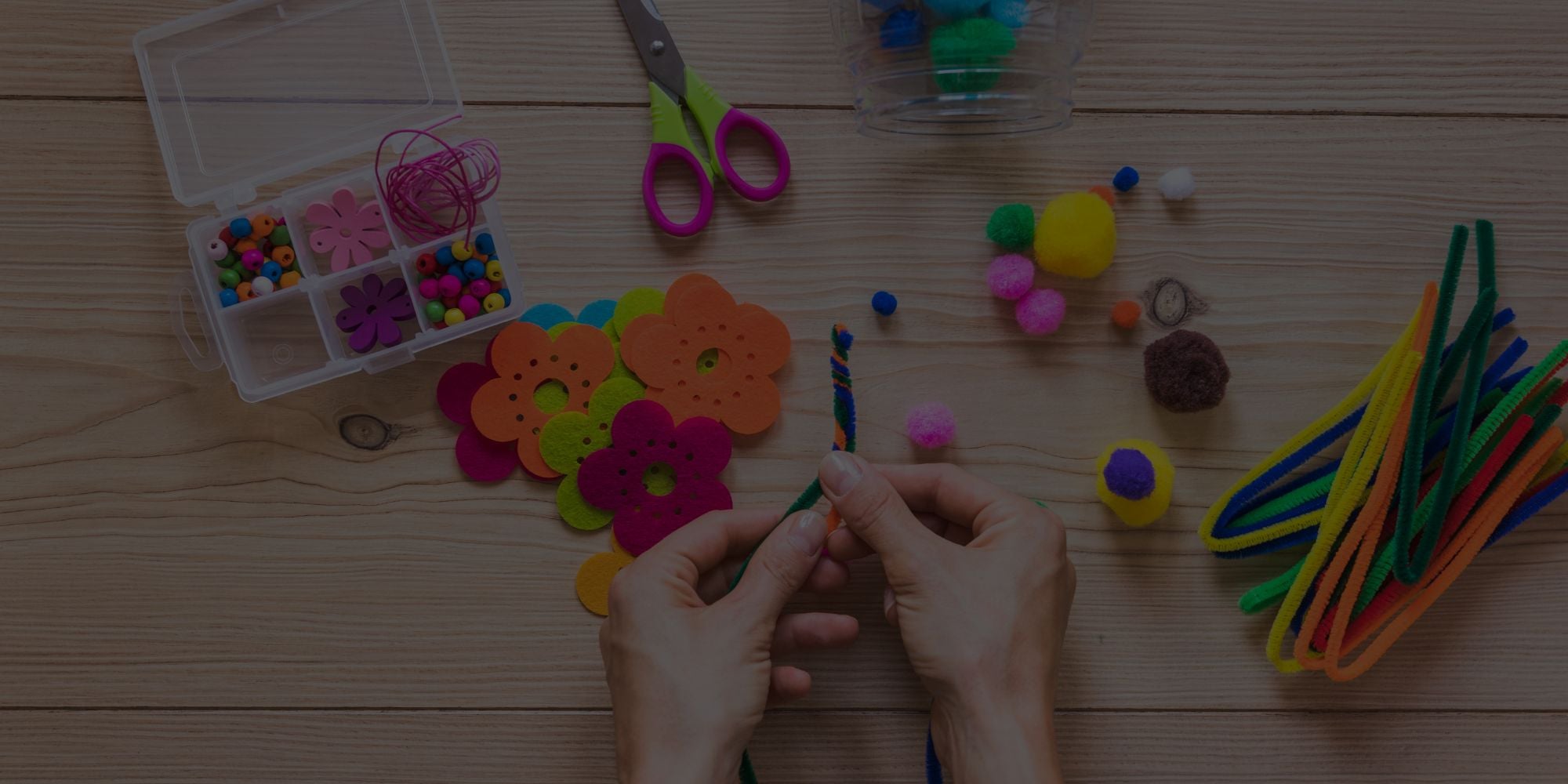 A top-down view of a person's hands crafting with colorful felt flower cutouts, pom-poms, pipe cleaners, and scissors on a light wood tabletop.