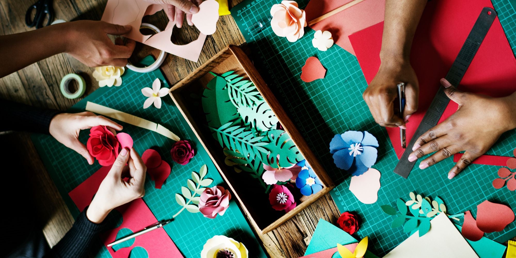 Hands working on paper crafts with cut-out shapes and flowers on a colorful cutting mat.