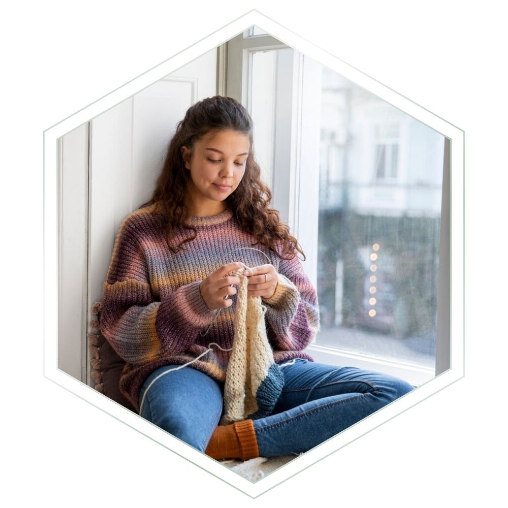 A woman sits by a window wearing a multicolored knit sweater and blue jeans, knitting with white yarn and metal needles. The photo is framed inside a white hexagonal border on a light gray background.