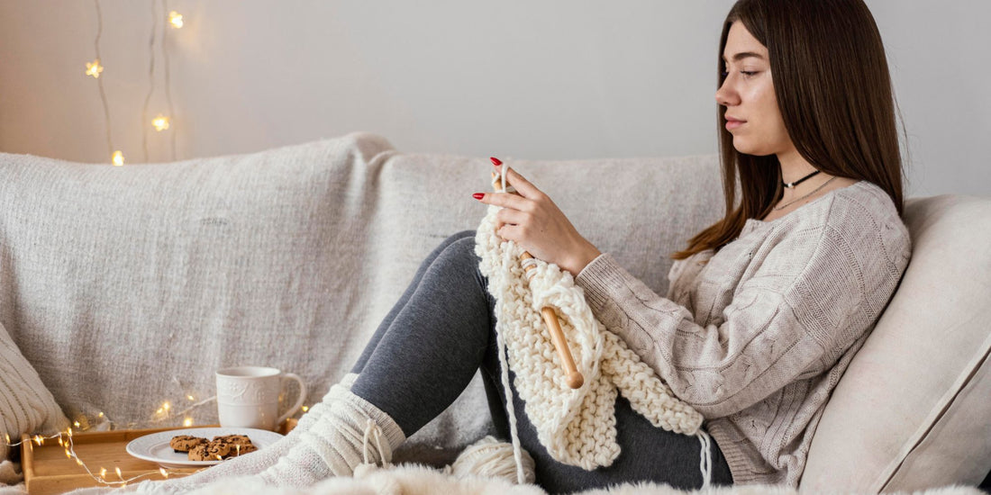 A woman sits on a beige couch, knitting with cream-colored yarn and wooden needles. She is wearing a beige knit sweater, dark gray leggings, and white socks, with a cup of coffee and cookies on a small table beside her.