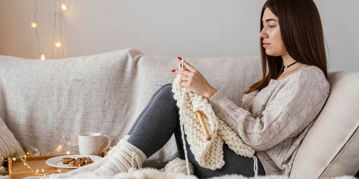 A woman sits on a beige couch, knitting with cream-colored yarn and wooden needles. She is wearing a beige knit sweater, dark gray leggings, and white socks, with a cup of coffee and cookies on a small table beside her.