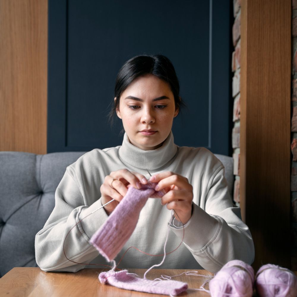 A woman sits at a wooden table, knitting with light purple yarn. She is wearing a light gray turtleneck sweater, with a gray tufted sofa behind her and a dark blue paneled wall in the background.