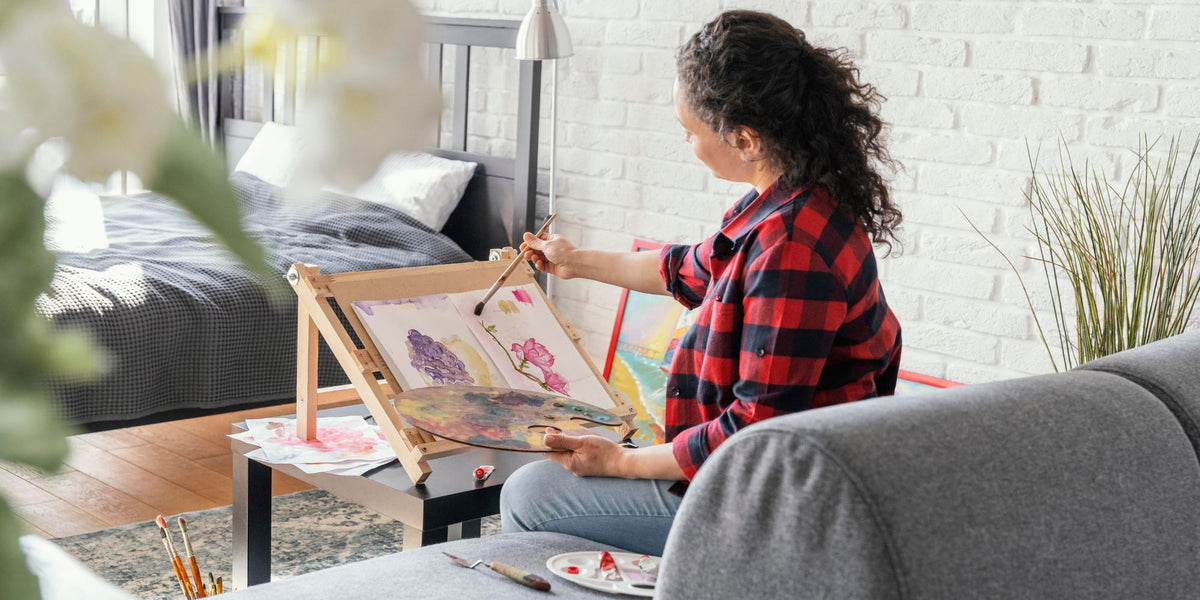 A woman with curly dark hair sits on a gray sofa in a bright bedroom, holding a paintbrush and painting on an easel. She wears a red and black plaid flannel shirt and light blue jeans.